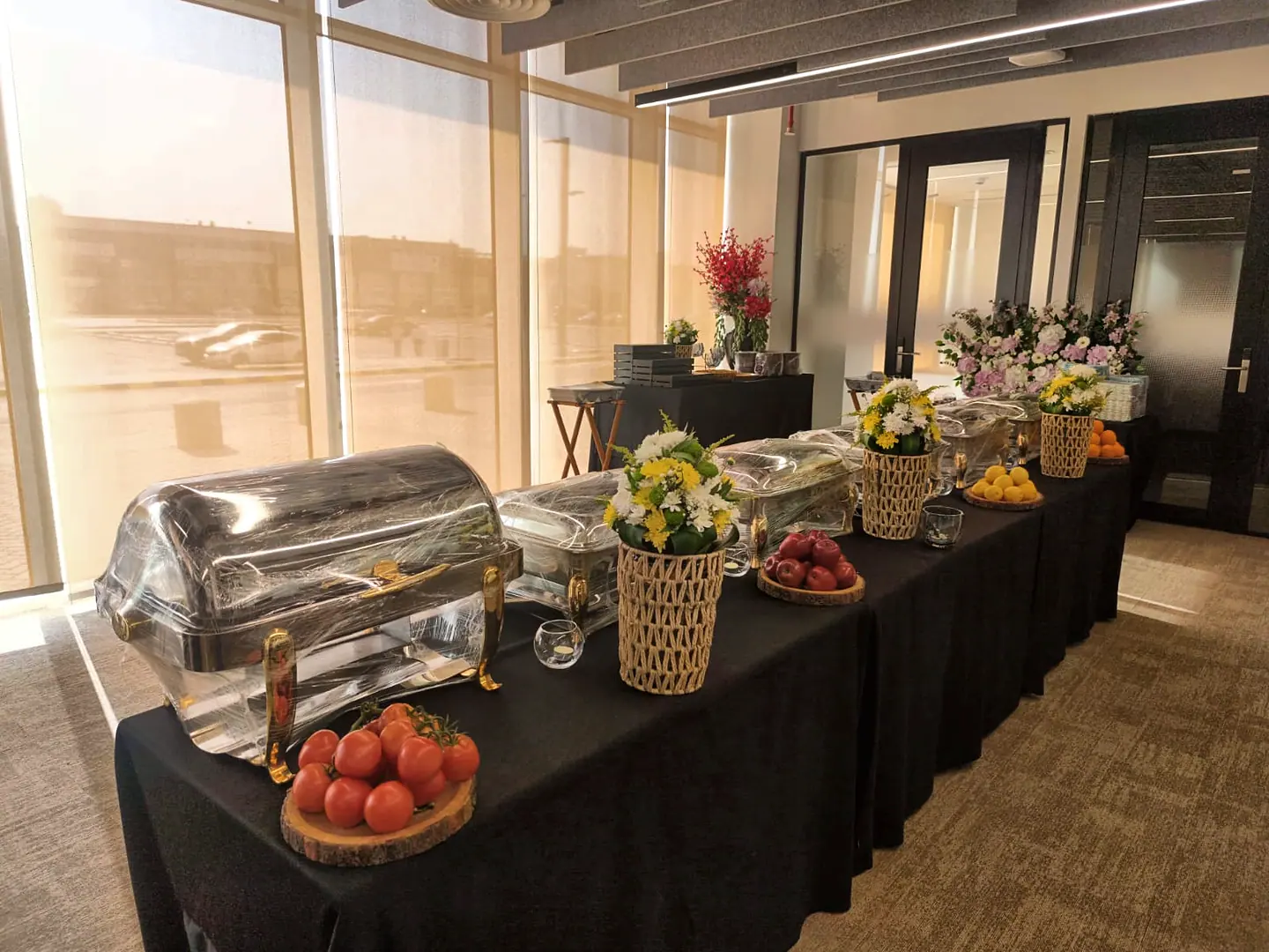 A buffet laid along black-draped tables: chafing dishes, tomatoes on wooden boards and baskets of yellow and white flowers